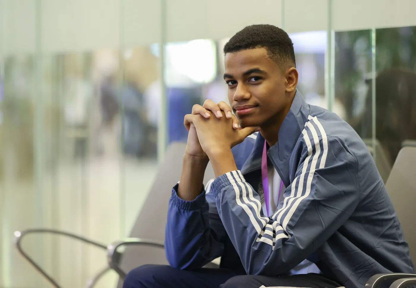 شاب يجلس في منطقة انتظار، يبتسم بأسلوب واثق مع يديه مشتبكتين أمامه. يرتدي سترة رياضية بأكمام طويلة ولون أزرق، مع شارة ملونة حول عنقه. في الخلفية، تظهر نوافذ زجاجية تعكس حركة الأشخاص.