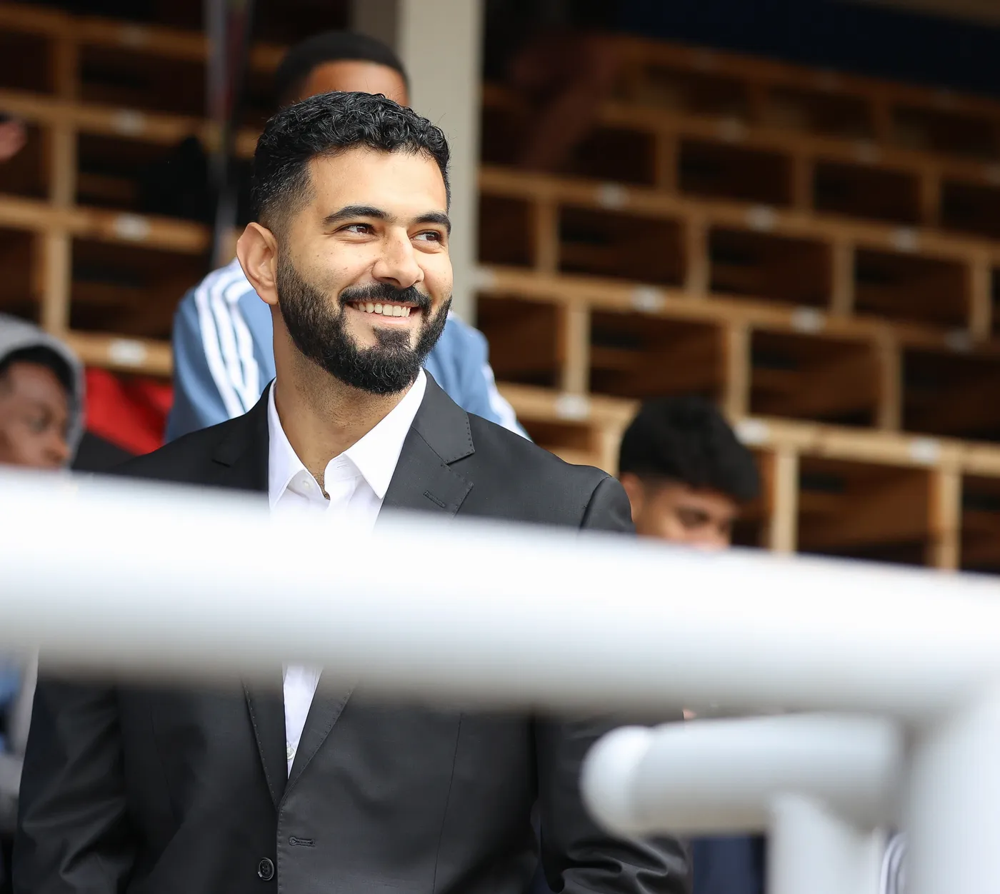 رجل يرتدي بدلة سوداء وقميصًا أبيض، مبتسمًا ويقف في مكان يشبه مدرجًا رياضيًا. خلفه يبدو بعض الأشخاص في ملابس غير رسمية، مما يضفي جوًا من التفاعل الاجتماعي.