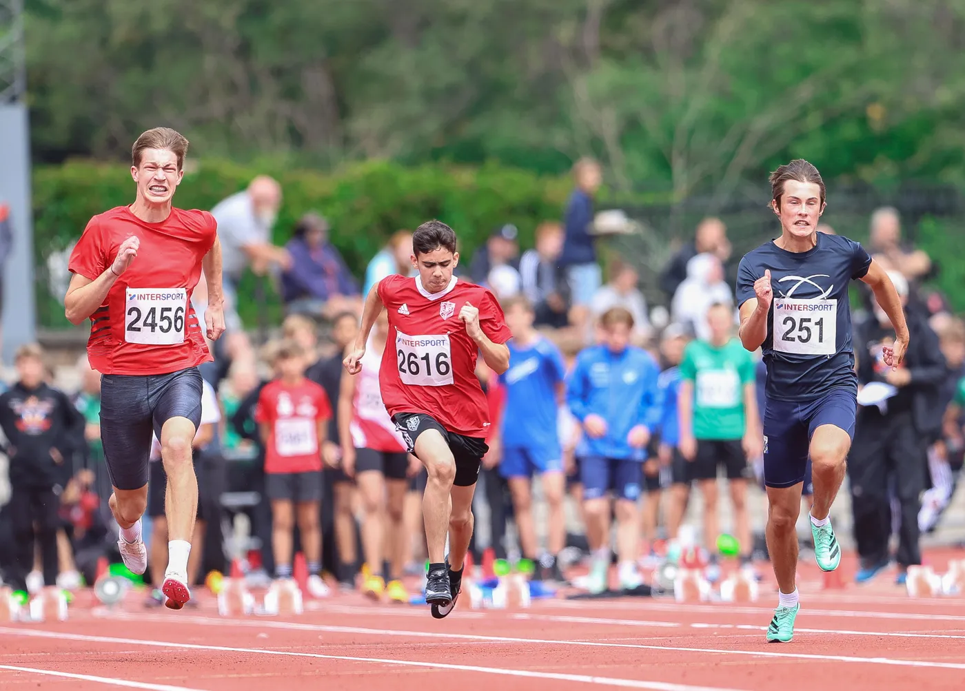 عداءان يرتديان قمصانًا حمراء يتنافسان في سباق، بينما يظهر عداء آخر يرتدي قميصًا أزرق في الخلفية. تشهد الحلبة الرياضية حضور مجموعة من المشجعين والأطفال، مع تركيز العدائين على الوصول إلى خط النهاية بأقصى سرعة.