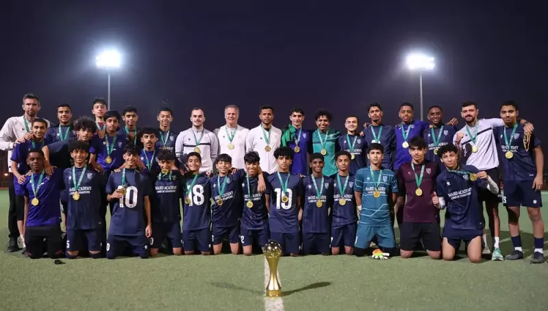 فريق لكرة القدم يتجمع بعد مباراة الفوز، مع كأس مميز في المقدمة. يرتدي اللاعبون قمصاناً زرقاء ويحملون ميداليات ذهبية، بينما يقف التدريب والجهاز الفني خلفهم تحت الأضواء.