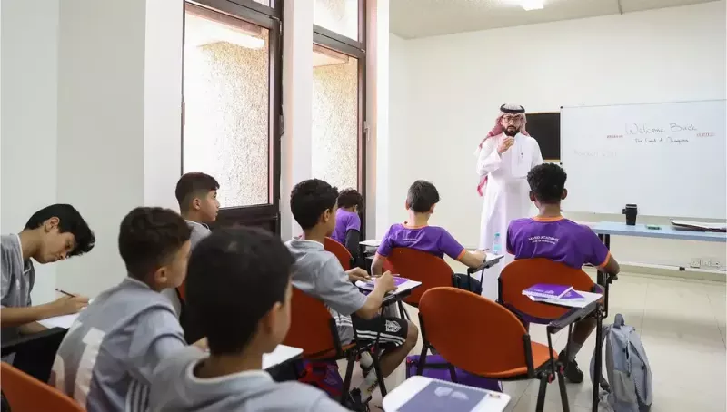 مدرّس يرتدي الثوب التقليدي يتحدث أمام مجموعة من الطلاب الذين يجلسون في صف دراسي. الطلاب يركزون في ملاحظاتهم بينما يظهر على السبورة عبارة 